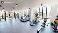 Located at 704 Rigsbee Ave in Durham, NC, this modern fitness center offers a well-appointed, single-floor workout space designed for a comprehensive exercise experience. The gym features a clean, contemporary aesthetic with crisp white walls, smooth high ceilings adorned with cylindrical pendant lighting, and durable dark rubberized flooring that spans the entire area.

Large floor-to-ceiling windows line the perimeter, flooding the space with abundant natural light and providing open views of the surrounding Durham streetscape. The bright, airy atmosphere enhances the overall workout environment, making the space feel expansive and inviting.

The fitness center is thoroughly equipped for both cardio and strength training. Cardio offerings include treadmills, an elliptical trainer, a stationary bike, and a rowing machine. Strength training equipment includes a multi-functional cable/smith machine rack, a dumbbell rack with a full range of free weights, adjustable benches, and plyometric boxes for functional training. A wall-mounted mirror provides visual feedback during workouts.

The layout is thoughtfully organized to maximize open floor space while keeping equipment accessible. The combination of modern equipment, generous natural light, and a clean design makes this fitness center a functional and motivating environment for residents or members in the heart of Durham.