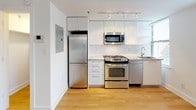 This single-floor unit features a clean, contemporary design characterized by white walls, crisp finishes, and warm honey-toned hardwood flooring that runs throughout the main living areas and bedroom. The open-plan layout connects the living space directly to the kitchen, creating an airy, cohesive environment well-suited for both everyday living and entertaining.

The kitchen is efficiently designed with sleek white cabinetry extending to the ceiling, stainless steel appliances including a gas range, built-in microwave, refrigerator, and dishwasher, complemented by a white countertop and a single basin sink. Track lighting overhead ensures the workspace is well-illuminated. A closet or utility area is accessible near the entry, providing additional storage.

The bedroom is generously proportioned, bathed in natural light through two large windows fitted with adjustable blinds that offer views of the surrounding urban landscape. The room connects directly to a bathroom featuring marble-style tiled walls, a soaking bathtub with an integrated shower, corner shelving, and a grab bar. The overall ambiance of the space is bright, minimal, and modern.
