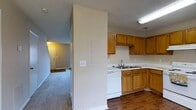 This two-floor residential space offers a practical and comfortable layout designed for everyday living. The kitchen is well-equipped with warm honey-toned wood cabinetry that provides generous storage, light-colored countertops, a double-basin sink, and white appliances including a dishwasher. The kitchen's hardwood flooring adds a classic touch and flows naturally into the adjoining areas. A structural dividing wall separates the kitchen from the adjacent living space, creating a sense of definition between the two areas while maintaining an open feel. The living and hallway areas feature plush carpeted flooring, contributing to a cozy atmosphere. The home's interior is finished with neutral, light-toned walls that keep the space feeling bright and airy. A textured ceiling is consistent throughout, and recessed lighting and flush-mount fixtures provide reliable illumination. The layout allows for easy movement between floors, with a staircase visible leading to the upper level. An entry door is visible at the end of the hallway, offering convenient access. Overall, the space presents a straightforward and livable floor plan suited for comfortable day-to-day use.
