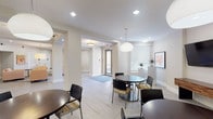 This well-appointed single-floor amenity space is designed to serve as a welcoming communal hub with a clean, contemporary aesthetic. Light-toned large-format tile flooring runs throughout, lending a cohesive and polished look to every area.

The open dining area features multiple round tables paired with a mix of sleek black and patterned seating, illuminated by striking globe pendant lights. Natural light filters through slatted blinds on the windows, creating a bright and inviting atmosphere. A wall-mounted flat-screen television and a rustic wood credenza anchor one end of the dining space.

Adjacent to the dining area is a comfortable lounge zone furnished with warm amber upholstered armchairs, a wood-topped drum coffee table, and coordinating side tables. Recessed lighting and an oversized drum pendant provide layered illumination, while framed abstract artwork adds pops of color to the neutral walls. A second flat-screen television on a wooden stand completes the media-ready setup.

A dedicated coffee and beverage station offers a built-in counter with dark cabinetry, a single-serve coffee maker, and supply organizers — ideal for daily resident use. Nearby, a spacious mail room is lined with banks of individual metal mailboxes on both walls, brightened by overhead flush-mount lighting and additional colorful artwork displayed along the corridor.
