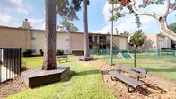 This single-floor residence combines thoughtful interior design with a welcoming outdoor environment. The building exterior is clad in classic brick, framed by towering mature trees and lush green lawns that create a serene, shaded setting. The communal outdoor area features benches, decorative landscaping, and open green space, providing a relaxed atmosphere for residents.

Inside, the interiors are finished with smooth, warm neutral-toned walls that lend a clean and airy feel throughout. The flooring features rich, dark concrete or stained hardwood, grounding the space with an earthy, industrial character. Recessed lighting illuminates the rooms with a soft, even glow.

The dining area is a standout feature, centered around a striking round marble-top table with a sculptural pedestal base, accompanied by woven rattan chairs atop a textured area rug. A large framed abstract artwork adorns the wall, adding an artistic focal point to the room. A sleek sliding barn door connects spaces with a functional yet stylish touch.

An adjacent room, accessible through a set of rustic vintage-style doors, reveals additional wall art and décor, hinting at a creatively appointed living or lounge area. Throughout the residence, the combination of curated artwork, natural materials, and warm neutral tones creates a cohesive and inviting ambiance.