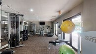 This fitness center is a comprehensive, single-floor workout facility designed to support a wide range of exercise routines. The space is finished with durable, textured carpeting throughout and features neutral gray walls that create a clean, focused atmosphere. Recessed lighting illuminates the room evenly, complemented by natural light streaming in through large glass doors and windows that open to an outdoor patio area, offering pleasant outdoor views of lush greenery.

The strength training zone is well-appointed, featuring a multi-station cable machine, a dedicated weight bench, and a full dumbbell rack with a wide range of hex weights neatly organized in ascending order. Floor-to-ceiling mirrors along one wall visually expand the space and allow users to monitor their form during workouts.

The cardio area includes a treadmill and an elliptical trainer, with a wall-mounted flat-screen television positioned for easy viewing during exercise sessions. A collection of stability and exercise balls in varying sizes adds versatility for core and flexibility training.

The fitness center also provides easy outdoor access through glass-panel doors, connecting the interior workout space to an exterior patio. The overall layout is efficient and thoughtfully organized, offering a fully functional environment suitable for both cardiovascular and strength-based fitness routines.