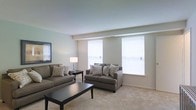 This single-floor space offers a clean, understated aesthetic defined by neutral tones and a welcoming, functional layout. The living area is a standout feature, showcasing wall-to-wall carpeting that adds warmth underfoot and a soft, comfortable feel throughout. Two large windows dressed with white horizontal blinds allow abundant natural light to fill the room, creating an airy and open atmosphere.

The living room is thoughtfully arranged with two gray upholstered sofas accented by decorative patterned throw pillows, centered around a dark rectangular coffee table. A matching side table with a simple table lamp provides additional ambient lighting, while a framed mirror on the wall adds depth and dimension to the space. The walls are finished in a soft, muted tone that complements the overall neutral color palette, contributing to a calm and cohesive interior environment.

The smooth ceiling and clean lines throughout the space reflect a straightforward, modern residential design that prioritizes comfort and practicality. The layout feels well-proportioned, offering ample room for both relaxation and everyday living. Overall, this space presents a tidy, move-in-ready environment suited for comfortable day-to-day use.