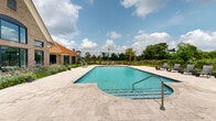 Located in Lake Charles, Louisiana, this outdoor pool facility offers a well-designed recreational space ideal for relaxation and leisure. The centerpiece is a generously sized swimming pool with clear blue water, surrounded by elegant travertine tile decking that extends throughout the area, providing a clean and polished aesthetic underfoot.

The pool area is framed by thoughtfully arranged landscaping, including ornamental grasses, tropical palms, neatly trimmed shrubs, and potted plants that add lush greenery and a sense of privacy. Lounge chairs are positioned along the pool's perimeter, offering comfortable spots to enjoy the open sky and scenic views of the surrounding Louisiana landscape.

A covered walkway flanked by brick columns connects the pool deck to the adjacent single-story clubhouse building, which features classic brick exterior walls, a distinctive copper-toned metal roof, and large windows that allow views into and out of the interior. The entrance to the clubhouse is accessed via sleek glass doors framed in dark metal. A covered porch near a secondary entrance includes a classic wooden bench and wall-mounted lantern lighting, adding a welcoming touch. The combination of durable brick construction, refined finishes, and abundant natural surroundings creates a polished and inviting outdoor environment.