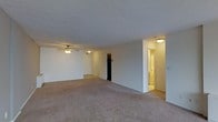 This single-floor space features an open, generously sized main living area finished with wall-to-wall neutral-toned carpeting that extends throughout, lending a cohesive and comfortable feel underfoot. The room is characterized by smooth, light-colored walls and a textured ceiling, contributing to a clean and understated aesthetic. A ceiling fan with an integrated light fixture is centrally mounted, providing both air circulation and ambient lighting to the space.

The layout opens toward multiple doorways, suggesting easy access to additional rooms. One opening reveals a darkened closet or storage area, while another leads to a warmly lit hallway or adjoining room, hinting at a bathroom or secondary space bathed in soft yellow light. The transition between areas is defined by subtle architectural framing, maintaining an open yet organized flow throughout the unit.

Overall, the space presents a functional and straightforward layout with neutral finishes that offer a versatile canvas. The even lighting, open floor plan, and accessible room connections make it a practical and adaptable living environment suitable for a variety of arrangements.