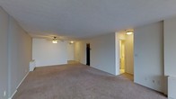This single-floor space features an open, generously sized main living area finished with wall-to-wall neutral-toned carpeting that extends throughout, lending a cohesive and comfortable feel underfoot. The room is characterized by smooth, light-colored walls and a textured ceiling, contributing to a clean and understated aesthetic. A ceiling fan with an integrated light fixture is centrally mounted, providing both air circulation and ambient lighting to the space.

The layout opens toward multiple doorways, suggesting easy access to additional rooms. One opening reveals a darkened closet or storage area, while another leads to a warmly lit hallway or adjoining room, hinting at a bathroom or secondary space bathed in soft yellow light. The transition between areas is defined by subtle architectural framing, maintaining an open yet organized flow throughout the unit.

Overall, the space presents a functional and straightforward layout with neutral finishes that offer a versatile canvas. The even lighting, open floor plan, and accessible room connections make it a practical and adaptable living environment suitable for a variety of arrangements.