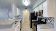 This single-floor space showcases a thoughtfully designed galley-style kitchen as a central highlight. The kitchen features crisp white shaker cabinetry lining both walls, providing ample upper and lower storage. Light gray speckled countertops run along each side, complemented by a stainless steel sink on one wall and a full suite of stainless steel appliances on the other, including a gas range, over-the-range microwave, refrigerator, and dishwasher. A tasteful mosaic tile backsplash adds a subtle decorative touch behind the range. Light wood-toned luxury vinyl flooring runs throughout, contributing to a clean, cohesive aesthetic. Soft blue-gray walls create a calm, neutral backdrop, while a flush-mount ceiling light provides bright overhead illumination. A doorway at the end of the kitchen leads into an adjoining room, suggesting an open and connected interior layout. The overall design balances functionality with modern finishes, resulting in a bright and practical living environment.
