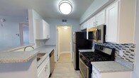 This single-floor space showcases a thoughtfully designed galley-style kitchen as a central highlight. The kitchen features crisp white shaker cabinetry lining both walls, providing ample upper and lower storage. Light gray speckled countertops run along each side, complemented by a stainless steel sink on one wall and a full suite of stainless steel appliances on the other, including a gas range, over-the-range microwave, refrigerator, and dishwasher. A tasteful mosaic tile backsplash adds a subtle decorative touch behind the range. Light wood-toned luxury vinyl flooring runs throughout, contributing to a clean, cohesive aesthetic. Soft blue-gray walls create a calm, neutral backdrop, while a flush-mount ceiling light provides bright overhead illumination. A doorway at the end of the kitchen leads into an adjoining room, suggesting an open and connected interior layout. The overall design balances functionality with modern finishes, resulting in a bright and practical living environment.