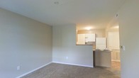 Located at 2591 Venturi House in Columbus, Ohio, this single-floor residence offers a clean, straightforward layout designed for comfortable everyday living. The living area features wall-to-wall carpeting in a neutral tone, providing a warm and quiet underfoot experience. Smooth, light-colored walls contribute to a bright and open ambiance throughout the space. A defining architectural feature is the pass-through kitchen, which connects seamlessly to the living area via a raised countertop peninsula. The kitchen is equipped with white cabinetry, a white refrigerator, and a microwave, all set against contrasting dark countertops that add visual interest. The overhead lighting in the kitchen provides focused illumination, keeping the cooking area well-lit. The open sightlines between the living and kitchen areas foster an interconnected feel, making the space functional for both relaxing and entertaining. White baseboards frame the perimeter of the living room, offering a clean, finished look. The overall design is practical and move-in ready, with a neutral color palette that provides a versatile backdrop for a variety of interior styles.