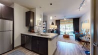 This contemporary single-floor residence is thoughtfully designed with an open-concept layout that seamlessly connects the kitchen, dining, and living areas. The kitchen is a standout feature, showcasing rich dark espresso cabinetry, granite countertops, stainless steel appliances including a refrigerator, microwave, and dishwasher, and a green tile backsplash. A peninsula with bar seating and elegant pendant lighting serves as a natural dividing point between the kitchen and living space. Wood-look plank flooring runs throughout the main living areas, adding warmth and continuity. The living room is bright and inviting, benefiting from large sliding glass doors that open to an outdoor patio and allow generous natural light to fill the space. A ceiling fan adds comfort to the area. The bedroom is generously sized with plush carpeting, a ceiling fan, and a large window framing outdoor views. The full bathroom is clean and modern, featuring dark vanity cabinetry with granite countertops, a glass-enclosed walk-in shower with tile surround, and wood-look flooring that mirrors the rest of the unit. The overall design balances contemporary aesthetics with functional living, creating a cohesive and comfortable environment throughout.
