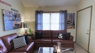 This two-story residence offers a welcoming living space anchored by a rich, dark brown leather sectional sofa that serves as the focal point of the room. The seating area is styled with contrasting throw pillows, including bold black cushions and a chevron-patterned accent pillow, adding visual interest and texture to the space. A glass-topped coffee table with a metallic frame sits at the center, complementing the leather furniture with a sleek, modern touch.

Natural light enters through a centrally positioned window dressed with sheer blue-gray grapetop curtains layered over horizontal blinds, lending a soft ambiance to the room. The neutral-toned walls provide a clean backdrop for decorative elements, including a vibrant cityscape wall art piece on one side and a classic table lamp on the opposite end. A distinctive green horse figurine adds a playful, artistic accent to the side table.

The overall atmosphere is casual yet curated, blending comfortable furnishings with thoughtful décor choices. The two-floor layout suggests additional living or sleeping space beyond the main living area, offering functional versatility throughout the home.