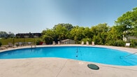 Located at 6516 N University St in Peoria, Illinois, this outdoor pool facility offers a well-appointed recreational space designed for relaxation and leisure. The centerpiece is a generously sized kidney-shaped swimming pool featuring vivid blue water, a clearly marked lap lane divider, and built-in entry steps with handrails for safe access.

Surrounding the pool is a broad, level concrete deck that provides ample room for lounging and movement. Multiple sling-style lounge chairs are positioned around the perimeter, offering plenty of spots to relax in the open air. The deck also features designated drainage channels to manage water runoff efficiently.

The pool area is fully enclosed by a black iron fence, ensuring both safety and security. A utility shed is situated at one end of the enclosure, providing convenient storage for pool equipment. An adjacent brick and shingle-clad building with a covered section adds architectural character to the space, and lounge furniture is arranged along its base for additional seating.

The entire area is framed by a lush backdrop of mature deciduous trees, creating a natural, park-like atmosphere. The setting benefits from abundant open-sky exposure, allowing for plentiful natural light throughout the day. The combination of thoughtful amenities, secure enclosure, and green surroundings makes this a comfortable and inviting outdoor recreational destination.