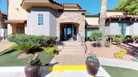 This single-story community space makes a strong first impression with its Mediterranean-inspired exterior, showcasing a striking stone archway entrance, stucco façade with a tile roof, and thoughtfully landscaped surroundings featuring desert-adapted plants, ornamental shrubs, and decorative oversized planters. A wide, accessible walkway leads visitors to the front entrance, framed by manicured greenery that adds a welcoming touch.

Inside, the space opens into a bright, expansive main area defined by soaring two-story ceilings that flood the room with natural light through large upper-level windows. Rich, warm-toned hardwood flooring runs throughout, anchoring the open layout with an inviting sense of warmth. Crown molding adds a refined architectural detail along the upper walls.

The interior is thoughtfully appointed with a central high-top seating area ideal for casual gatherings or meetings, complemented by a wall-mounted flat-screen television and a dedicated work or service counter with dark cabinetry. Tall decorative trees placed throughout the space add a natural, lively element to the interior. A ceiling fan assists with comfort, while French doors at the rear of the space provide a direct connection to an outdoor area, allowing ample natural light to pour in and creating a seamless indoor-outdoor flow.