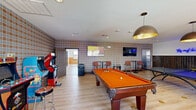 This expansive single-floor amenity center spans a generous footprint and is thoughtfully designed to provide a diverse range of recreational and social spaces. The game room serves as a central highlight, featuring distinctive plaid-patterned wallpaper, light hardwood flooring, and an array of entertainment options including a pool table, ping-pong table, and classic arcade cabinets such as an NBA and Ridge Racer machine. Overhead, large bronze pendant lights add a stylish touch, while wall-mounted TVs and bar-height seating areas along the perimeter create an ideal social atmosphere. Adjacent lounge areas offer comfortable booth-style seating lined along oversized windows that invite abundant natural light and views of the surrounding neighborhood and pool area. Bold navy toile wallpaper and wood wainscoting lend a distinctive, curated character to these spaces. The fitness center is enclosed by sleek floor-to-ceiling glass walls and sliding doors, offering full visibility into the bright, well-ventilated room. Inside, multiple treadmills, stationary bikes, and strength-training equipment are arranged across light hardwood floors, complemented by ceiling fans, wall-mounted TVs, and mirrors that enhance the sense of openness. A connecting corridor with water fountains and greenery ties the spaces together seamlessly.