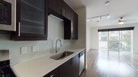 This single-floor unit showcases a contemporary design with a cohesive palette of dark espresso cabinetry, light countertops, and warm wood-style flooring that flows throughout the main living areas. The galley-style kitchen is a standout feature, outfitted with rich dark cabinetry — including glass-front upper cabinets — a subtle mosaic tile backsplash, stainless steel appliances including a gas range, microwave, refrigerator, and dishwasher, as well as a stainless steel undermount sink with a modern pull-down faucet. Track lighting overhead provides functional illumination across the full length of the kitchen. The open living area is bright and airy, bathed in natural light streaming through large sliding glass doors that lead to a private balcony with views of the surrounding neighborhood. A ceiling fan adds comfort to this versatile space. The bathroom features dark cabinetry with ample drawer storage, a white undermount sink set into a clean countertop, large-format tile flooring, and a generously sized mirror. A sliding barn-style door adds a modern touch to the bathroom entry. The bedroom, accessible from the main living space, offers hardwood-style flooring, clean white walls, and its own sliding glass door opening to the balcony, creating a bright and connected retreat.