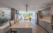 Located at 1505 Commonwealth Ave in Boston, MA, this modern community lounge is a thoughtfully designed shared space that combines functionality with contemporary style. The open-plan layout flows seamlessly from a fully equipped kitchen area into distinct lounge and social zones, all unified by warm wood-look flooring and clean white ceilings with recessed linear LED lighting.

The kitchen is a standout feature, boasting sleek grey cabinetry, expansive white quartz countertops, a large center island with an integrated sink and dishwasher, and stainless steel appliances including a full-size refrigerator. A striking pendant light fixture composed of clustered mason jar-style lamps hangs above the island, adding an artisan touch to the modern aesthetic.

The lounge area offers multiple seating arrangements to suit different social settings. Bold yellow accent chairs near the floor-to-ceiling windows create a vibrant, energetic nook filled with natural light and views of the greenery outside. A separate sitting area features tufted leather sofas and armchairs arranged around a sculptural cloud-shaped table, complemented by eclectic décor including a gold antler wall mount and a mannequin floor lamp.

An eye-catching patchwork tile-inspired accent wall adds texture and artistic depth to the space, making it a truly distinctive and welcoming environment.