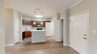 Located at 5462 Whittlesey Blvd in Columbus, Georgia, this single-floor residence offers a well-designed layout that balances functionality with comfortable living. The open-concept kitchen serves as the heart of the home, showcasing rich dark wood cabinetry, white countertops, and a full suite of black appliances including an electric range, microwave, and refrigerator. A central island with pendant lighting above provides additional prep space and a natural gathering point. Wood-style luxury vinyl flooring flows throughout the main living areas, lending a clean and cohesive aesthetic. Crown molding adds a refined architectural touch in key areas of the home. The living area is generously proportioned, featuring a ceiling fan and a large window that invites natural light and offers pleasant views of the surrounding greenery. Bedrooms are finished with soft wall-to-wall carpet and include ceiling fans, with windows framing lush outdoor foliage. Ample closet storage is present throughout. The bathroom is well-appointed with a soaking tub and shower combination, a rich wood vanity with a light countertop, and wood-style flooring that ties the space together. The overall ambiance is warm, neutral, and move-in ready.
