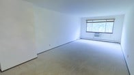This single-floor space offers a clean and functional layout designed for comfortable everyday living. The main living area is generously proportioned, featuring neutral wall-to-wall carpeting that adds warmth and a sense of openness to the room. Bright white walls create a clean, airy backdrop that makes the space feel fresh and inviting.

A large window serves as a focal point of the living area, allowing abundant natural light to flood the room and offering views of lush greenery outside. The window is fitted with a roll-up shade for adjustable light control and privacy. A wall-mounted air conditioning unit positioned beneath the window ensures year-round climate comfort.

The baseboard heating system runs along the base of the walls, providing efficient and consistent warmth throughout. The room's straightforward, open design offers flexibility for a variety of furniture arrangements and décor styles. Overall, the space presents a simple yet functional environment with practical amenities, making it well-suited for those seeking a low-maintenance, move-in ready home with natural light and outdoor views.