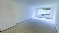 This single-floor space offers a clean and functional layout designed for comfortable everyday living. The main living area is generously proportioned, featuring neutral wall-to-wall carpeting that adds warmth and a sense of openness to the room. Bright white walls create a clean, airy backdrop that makes the space feel fresh and inviting.

A large window serves as a focal point of the living area, allowing abundant natural light to flood the room and offering views of lush greenery outside. The window is fitted with a roll-up shade for adjustable light control and privacy. A wall-mounted air conditioning unit positioned beneath the window ensures year-round climate comfort.

The baseboard heating system runs along the base of the walls, providing efficient and consistent warmth throughout. The room's straightforward, open design offers flexibility for a variety of furniture arrangements and décor styles. Overall, the space presents a simple yet functional environment with practical amenities, making it well-suited for those seeking a low-maintenance, move-in ready home with natural light and outdoor views.