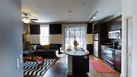This single-floor residence offers a thoughtfully designed open-concept layout that seamlessly integrates the kitchen, dining, and living areas into one cohesive and functional space. The kitchen is a standout feature, boasting dark cabinetry, granite countertops, stainless steel appliances including a refrigerator and dishwasher, and a peninsula island with bar seating — ideal for casual dining or entertaining. Pendant lighting above the island adds a refined touch, while track lighting overhead ensures the space is well-lit throughout. Rich hardwood-style flooring flows throughout the main living area, adding warmth and visual continuity. The living area is generously sized and anchored by a large sectional sofa, with bold décor accents including a chevron area rug and vibrant throw pillows that contribute to the space's contemporary aesthetic. A ceiling fan provides added comfort, while large sliding glass doors and expansive windows dressed in dramatic striped curtains flood the space with natural light and offer direct access to an outdoor area. The overall ambiance is modern and stylish, combining dark, contrasting tones with warm textures to create an inviting and well-appointed interior.
