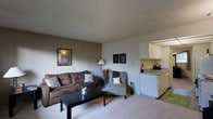 This single-floor residence offers a comfortable and efficiently designed layout that makes excellent use of its space. The living area serves as the heart of the home, featuring plush wall-to-wall carpeting underfoot and neutral, warm-toned walls that create a welcoming ambiance. A textured ceiling adds subtle visual interest overhead. The living room provides ample space for a full sofa, accent chair, and coffee table arrangement, with room for decorative touches such as wall art and floor lamps that enhance the cozy atmosphere. An open pass-through connects the living area directly to the kitchen, promoting an airy, open-concept feel. The kitchen is well-appointed with white cabinetry offering generous storage, a single-basin sink, and tile flooring that contrasts neatly with the carpeted living space. The cabinetry extends to the upper walls, maximizing vertical storage. The overall design emphasizes functionality and simplicity, with a clean, understated aesthetic defined by its neutral color palette. Natural light filters into the space, contributing to a bright and inviting environment. This single-level layout offers straightforward, accessible living with a cohesive flow between the main living and kitchen areas.