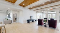 Located on Mulberry Blvd in Port Wentworth, GA, this single-story community clubhouse offers a generously sized, thoughtfully designed gathering space with a range of amenities. The main lounge is an open, airy hub defined by striking wood-paneled beam ceilings, recessed lighting, and light-toned tile flooring that flows throughout. Floor-to-ceiling black-framed glass walls and doors frame direct views of the outdoor pool and patio area, flooding the interior with abundant natural light and creating a seamless indoor-outdoor connection. A shuffleboard table and plush jewel-toned seating provide a sophisticated game and social area, complemented by a wet bar and a wall-mounted television. A dedicated dining lounge features modular booth-style seating, pedestal tables, and a series of pendant lights beneath serene coastal artwork — ideal for casual gatherings. A private conference room is finished with deep green wainscoting, a long conference table with leather chairs, a wall-mounted display, and a decorative pendant chandelier, suited for professional meetings. An additional flexible room with large draped windows and ample natural light serves as a versatile workspace or reading area. Throughout, the design maintains a clean, contemporary aesthetic with warm architectural accents and curated furnishings.
