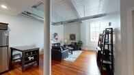 This single-floor urban loft blends industrial character with modern finishes throughout. The expansive living area immediately impresses with its dramatically high ceilings, exposed HVAC ductwork, and painted wood-plank ceiling details that nod to the building's heritage. Rich hardwood floors run throughout, anchoring the open, airy layout and complementing the neutral gray walls.

The kitchen is a standout, outfitted with dark espresso shaker cabinets, granite countertops, and a full suite of stainless steel appliances including a refrigerator, range, and microwave. A generous center island with seating provides additional prep space and casual dining. Pendant lighting overhead adds warmth to the functional layout.

The bedroom is generously proportioned, with the same soaring ceilings found throughout the unit, and benefits from access to a walk-in closet and a dedicated bathroom. The en-suite bathroom features a dual-sink vanity with granite countertops, white cabinetry, classic mosaic tile flooring, and a glass-enclosed shower.

A dedicated laundry room houses a stacked washer and dryer alongside a fold-out utility surface, adding everyday convenience. Large windows allow natural light to filter through the living spaces, enhancing the loft's open, industrial-chic ambiance.