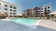 Located at 5085 Main St in Trumbull, Connecticut, this outdoor amenity space centers around a generously sized swimming pool with crystal-clear water, set against a striking multi-story building complex. The architectural design blends classic New England influences with modern construction, showcasing a combination of stone masonry, deep red and white exterior facades, pitched gabled rooflines, and large multi-pane windows that lend the property a refined, contemporary character.

The pool deck is finished with large-format stone-patterned pavers that extend broadly around all sides of the pool, providing ample space for relaxation and movement. Rows of cushioned lounge chairs and chaise lounges line the perimeter, offering plentiful sunbathing opportunities. Several shaded seating areas with large market umbrellas and outdoor dining chairs are positioned throughout the deck, ideal for socializing or enjoying the open-air setting.

A covered entry or pavilion area with a copper-accented roof adds architectural detail along the building's pool-facing facade, which features prominent natural stone cladding. The entire pool area is enclosed by a clean wrought-iron fence, ensuring privacy while maintaining unobstructed sightlines to the surrounding buildings and open sky. The space benefits from abundant natural sunlight, making it a well-appointed outdoor retreat.