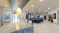 This single-floor clubhouse and business center combines refined style with practical functionality, offering residents an upscale environment for both social gatherings and productive work. The space is anchored by a stunning marble-topped bar or counter, illuminated by warm, oversized pendant lights with gold accents that add a touch of elegance to the open layout. Light-toned porcelain tile flooring flows seamlessly throughout, enhancing the bright and airy atmosphere created by abundant recessed lighting and generous natural light from large windows and glass doors. A spacious dining and lounge area features a long conference-style table surrounded by contemporary blue and neutral upholstered chairs, ideal for meetings or community events. A wall-mounted flat-screen TV serves as a focal point for the lounge zone, which is further accented by tasteful artwork and decorative elements. Soft, neutral tones on the walls complement the blue drapery and furnishings, creating a cohesive and polished aesthetic. The open floor plan allows for flexible use of the space, accommodating both intimate gatherings and larger group events with ease. Overall, this clubhouse and business center delivers a professional yet inviting atmosphere designed to serve a wide range of community needs.
