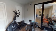 This fitness center is a compact yet fully functional workout facility designed to support a variety of exercise routines. The space features durable wood-look flooring throughout, neutral-toned walls, and adequate overhead lighting to create a practical and focused environment for physical activity.

The main workout area is outfitted with several pieces of cardio equipment, including an elliptical trainer and stationary spin bikes, all positioned for easy use. A dumbbell rack with multiple weight options lines one wall, accompanied by a weight bench, catering to strength training needs. A large wall-mounted flat-screen television is positioned prominently for entertainment during workouts, with seating available nearby.

Near the glass entry door, a tiered medicine ball rack holds a range of weighted balls, offering additional options for functional fitness training. The glass-paneled entry door allows natural light to filter in and provides views of the surrounding outdoor area, contributing to an open and inviting atmosphere.

Interior glass-paned doors with bold black frames connect the fitness center to an adjacent common area, enhancing visibility and the sense of connectivity throughout the facility. Two additional interior doors provide access to separate spaces within the center. Overall, this fitness center delivers a well-rounded, amenity-rich environment suited for both cardio and strength-based workouts.