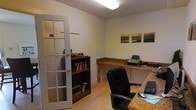 This single-floor office space offers a clean, functional environment designed for productivity and professional use. The room is accessed through a classic French door with multi-pane glass panels, adding a refined architectural touch while allowing light to pass between spaces. The interior features warm light wood flooring that contrasts neatly with the neutral-toned walls and a textured ceiling with flush-mount lighting that provides even, practical illumination throughout. A generous L-shaped desk with a granite-style surface wraps around one corner of the room, offering ample workspace for computing, writing, and office equipment. A dark wood bookcase stands nearby, providing convenient storage for reference materials and books. The walls are kept simple and uncluttered, adorned with a small gallery of framed coastal landscape artwork that adds a calm, professional ambiance to the space. The overall layout is efficient and well-organized, making the most of the available footprint. With its warm tones, thoughtful built-ins, and quiet atmosphere, this office space is well-suited for daily business operations, focused study, or administrative tasks.