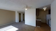 This single-floor unit is laid out in a compact, functional configuration that makes efficient use of its space. The living area is the largest zone in the unit, featuring carpet flooring and natural light streaming in through a window. A ceiling fan with a built-in light fixture is centered overhead, complementing the textured popcorn ceiling. The open layout connects the living area directly to the kitchen, creating a seamless flow between the two spaces.

The kitchen is positioned along one side of the unit and features dark wood cabinetry, a sink with under-cabinet storage, and a freestanding range with an oven. The kitchen flooring transitions to a wood-style surface, visually distinguishing it from the carpeted living area. Adjacent to the kitchen is a bathroom with basic fixtures and a separate enclosed area.

A notable feature of this unit is the generous closet storage, with two side-by-side sliding-door closets visible from the living area, each offering substantial hanging and shelving space. The walls are painted in neutral tones throughout, contributing to a clean and straightforward interior aesthetic. The overall layout is practical, with clearly defined functional zones within a modest footprint.