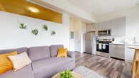Located on the third floor at 174 E Long St in downtown Columbus, Ohio, this one-bedroom, one-bathroom unit offers a thoughtfully designed layout that maximizes every square foot of its compact footprint. The open-plan living and kitchen area serves as the heart of the space, where hardwood-style flooring runs throughout, creating a cohesive and modern aesthetic. Large black-framed windows in the kitchen and living area flood the space with natural light and offer views of the surrounding Columbus cityscape.

The kitchen is well-appointed with stainless steel appliances, including a full-size refrigerator, electric range, over-the-range microwave, and dishwasher. Light gray cabinetry extends to the ceiling, maximizing storage, while white countertops keep the look clean and functional. A small dining area adjacent to the kitchen windows provides an ideal spot for casual meals.

The living room features a wall-mounted mini-split A/C unit and a distinctive golden-yellow accent soffit that adds a warm visual element to the otherwise bright, white walls. A hallway with a barn-style door leads to the private bedroom, which includes its own wall-unit A/C, a narrow clerestory window, and warm hardwood-style flooring topped with a colorful area rug. The bathroom is outfitted with a gray vanity, framed mirror, and a shower/tub combination.
