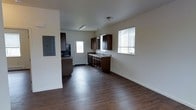 This single-floor residence offers a practical and functional layout designed for comfortable everyday living. The open main living area features rich, dark hardwood-style flooring that runs throughout, creating a cohesive and grounded aesthetic. Crisp white walls provide a clean contrast and help reflect light throughout the space.

The kitchen is integrated into the main living area and is equipped with dark espresso-toned cabinetry offering ample storage, complemented by a countertop sink and built-in appliances. A pendant light fixture hangs above, adding a subtle decorative touch. A window near the kitchen allows natural light to filter in, brightening the workspace.

An electrical panel is conveniently recessed into the wall in the central area of the home, keeping utilities accessible. A doorway from the main living space leads to additional rooms, suggesting a well-connected floor plan. The ceiling features a flush-mount light fixture that provides consistent overhead illumination throughout the main area.

Overall, the space presents a straightforward, no-frills design with functional finishes and a neutral color palette, making it a versatile and adaptable living environment suitable for a variety of needs.