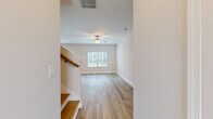 This two-story home offers six bedrooms and two bathrooms across two well-proportioned floors. The ground floor hosts the main living and kitchen areas, while the upper floor provides additional bedroom space and the primary suite.

The kitchen is a standout feature, equipped with white shaker-style cabinetry, a subway tile backsplash, stainless steel appliances, recessed lighting, and a central island with a granite countertop and built-in sink. Light wood-style flooring flows throughout the kitchen and living areas, contributing to a bright, cohesive aesthetic.

The living room is open and airy, featuring a ceiling fan, neutral white walls, and a window that allows natural light to fill the space. A staircase with a wood-railed banister connects the two floors.

The bedrooms are finished with plush carpet and ceiling fans, with windows providing natural light and exterior views. The primary bedroom includes access to a bathroom and a dedicated walk-in closet outfitted with wire shelving. The bathrooms are clean and functional, with one featuring a full tub-shower combination with chrome fixtures and wood-style flooring throughout.