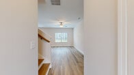 This two-story home offers six bedrooms and two bathrooms across two well-proportioned floors. The ground floor hosts the main living and kitchen areas, while the upper floor provides additional bedroom space and the primary suite.

The kitchen is a standout feature, equipped with white shaker-style cabinetry, a subway tile backsplash, stainless steel appliances, recessed lighting, and a central island with a granite countertop and built-in sink. Light wood-style flooring flows throughout the kitchen and living areas, contributing to a bright, cohesive aesthetic.

The living room is open and airy, featuring a ceiling fan, neutral white walls, and a window that allows natural light to fill the space. A staircase with a wood-railed banister connects the two floors.

The bedrooms are finished with plush carpet and ceiling fans, with windows providing natural light and exterior views. The primary bedroom includes access to a bathroom and a dedicated walk-in closet outfitted with wire shelving. The bathrooms are clean and functional, with one featuring a full tub-shower combination with chrome fixtures and wood-style flooring throughout.