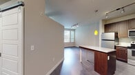 This well-appointed single-floor residence offers a thoughtfully designed layout that balances modern aesthetics with functional living. The open-concept kitchen is a standout feature, showcasing rich dark-wood cabinetry that extends to the ceiling, stainless steel appliances including a refrigerator, range, microwave, and dishwasher, a subway tile backsplash, and a granite-topped island with warm pendant lighting overhead. Track lighting further illuminates the space, adding a contemporary touch.

The living and dining area flows seamlessly from the kitchen, featuring dark hardwood-style flooring and a wall of windows fitted with horizontal blinds that invite generous natural light. A private exterior door with sidelight windows provides direct outdoor access, enhancing connectivity to the outside.

The bedrooms offer plush carpeting underfoot, each benefiting from natural light through blinds-fitted windows with views of surrounding greenery. One bedroom features a ceiling fan for added comfort. The bathroom is clean and functional, equipped with a soaking tub and shower combination, wood-style vinyl flooring, and neutral wall tones. Throughout the residence, soft neutral paint, clean white baseboards, and consistent finishes create a cohesive and inviting atmosphere.