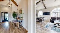This single-floor community clubhouse blends classic architectural charm with modern amenities. The space is immediately distinguished by its soaring vaulted ceilings adorned with rustic exposed wood beams, complemented by striking arched windows that flood every area with natural light and offer views of the surrounding neighborhood.

The main lounge area serves as the social heart of the space, featuring plush reclining seating arranged around a central coffee table atop a vibrant blue area rug. A wall-mounted television and generous window seating create an inviting atmosphere for both relaxation and community gatherings. An adjacent dining area with a round table and cushioned chairs provides additional seating, accented by wainscoting and continued arched window detailing.

A well-appointed kitchen offers rich, dark wood cabinetry with granite-style countertops, a classic white subway tile backsplash, and a full suite of stainless steel appliances including a gas range, microwave, refrigerator, and dishwasher. A compact counter with bar stools near the entry provides a casual spot for a quick bite.

The fitness center is spacious and well-lit, equipped with treadmills, ellipticals, a stair climber, free weights, and a weight bench. Motivational wall graphics, a water fountain, and a mirrored wall complete this energizing workout environment.