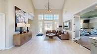 Located at 5555 W 6th St in Lawrence, Kansas, this single-floor space offers a thoughtfully designed layout comprising a living room, exercise room, and office. The living room is a standout centerpiece, boasting soaring two-story ceilings and an expansive wall of floor-to-ceiling windows and French doors that flood the space with abundant natural light and provide views of the surrounding outdoors. Light-toned large-format tile flooring, warm leather seating, a sculptural chandelier, and bold artwork contribute to a sophisticated yet welcoming ambiance. The office area features the same light tile flooring, bright windows, and open shelving with warm wood tones, providing a functional and organized workspace. The exercise room is well-equipped with cardio machines including treadmills and ellipticals, a cable weight machine, free weights, and a wall of mirrors that visually expand the space. A wall-mounted television, recessed lighting, and a distinctive tray ceiling with sage green accent tones give the fitness area a polished, purpose-built feel. Durable carpet flooring and roller shades on the windows complete the practical design. Together, these three spaces form a cohesive, multi-functional environment ideal for both relaxation and an active lifestyle.