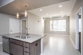This single-floor unit showcases a thoughtfully designed open-concept layout that seamlessly connects the kitchen and living area. The kitchen serves as a central focal point, featuring a large island with a double basin sink, quartz-style countertops, rich dark wood cabinetry extending to the ceiling, and a full suite of stainless steel appliances including a French door refrigerator, range, and dishwasher. Pendant lighting and a decorative geometric-patterned ceiling accent add visual interest above the island. Exposed industrial-style ductwork runs through the kitchen and bedroom areas, lending a modern, loft-inspired character to the space. Light tile flooring flows throughout the main living areas, while the bedroom features plush carpeting and a spacious walk-in closet with wire shelving. The living area benefits from natural light through large windows with blinds. Two bathrooms are finished with light tile flooring, soaking tubs with shower combinations, and dark wood cabinetry for storage. A dedicated in-unit laundry closet houses a full-size front-loading washer and dryer with overhead shelving, adding everyday practicality to this well-appointed home.