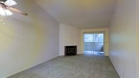 This single-floor residence offers a welcoming and functional layout designed for comfortable everyday living. The living area serves as the heart of the home, immediately drawing attention with its vaulted ceiling that adds a sense of openness and airiness to the space. Soft, neutral-toned wall-to-wall carpeting runs underfoot, contributing to a warm and understated ambiance throughout.

A classic corner fireplace with a traditional surround serves as a natural focal point, providing both a functional heating element and a charming architectural detail. The clean, white walls offer a bright and versatile backdrop, allowing for a wide range of interior styling possibilities.

One of the standout features of the living area is a full-width sliding glass door dressed with vertical blinds, which floods the room with generous natural light and provides direct access to an outdoor area. The view through the glass reveals lush greenery, enhancing the sense of connection to nature from within the home.

The overall design is clean and straightforward, with a neutral palette and minimal ornamentation that lends itself well to personalization. The single-floor layout ensures easy accessibility and flow throughout the space, making it a practical and inviting place to call home.
