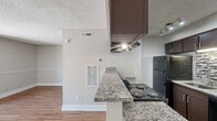 Located at 2320 S Conway Rd in Orlando, FL, this efficiently designed single-floor unit offers a thoughtful layout that maximizes its intimate footprint. The combined kitchen and living area forms the heart of the space, where warm wood-look flooring runs throughout, creating a cohesive and inviting atmosphere. The kitchen is well-appointed with dark espresso cabinetry, granite-style countertops, a full suite of black appliances including a refrigerator and electric range with overhead ventilation, and a tasteful tile backsplash accent. A peninsula counter with overhead cabinets separates the kitchen from the living area, providing both a functional workspace and a visual divider. Track lighting illuminates the kitchen corridor, adding a modern touch. The open living area is bright and airy, featuring neutral two-tone painted walls with chair rail detailing, crisp white baseboards, and a textured ceiling. A sliding glass door floods the space with natural light and opens directly onto an outdoor balcony with exterior views. The bathroom is clean and functional, featuring a full tub-shower combination with white tile surround and chrome fixtures. Overall, the unit delivers a practical and well-finished living environment.