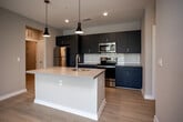Located at 906 Waterfront Dr in Knoxville, Tennessee, this single-floor residence combines modern design with functional living. The open-concept kitchen serves as a focal point, showcasing bold slate-blue cabinetry that extends to the ceiling, stainless steel appliances including a refrigerator, microwave, and range, a white subway tile backsplash, and a generous center island with a light quartz countertop and undermount sink. Stylish pendant lighting adds a contemporary touch overhead. Light-toned hardwood-style flooring runs throughout the main living areas, contributing to a bright and cohesive aesthetic. The living area connects seamlessly to a private balcony through sliding glass doors, offering pleasant outdoor views of the surrounding landscape and greenery. The bedrooms feature plush carpeting and receive natural light through well-placed windows. Closet storage is well-considered, with wire-rack shelving systems in multiple closets, including a dedicated walk-in option. The bathroom is finished with a wide single-sink vanity, a large mirror with overhead lighting, and a clean walk-in shower with glass enclosure. Throughout the space, neutral warm-gray walls, white trim, and recessed lighting create a polished and move-in-ready environment.