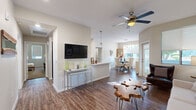 This single-floor residence offers a thoughtfully designed open-concept layout that seamlessly connects the living, dining, and kitchen areas. The living room is anchored by rich wood-look flooring, a ceiling fan, and a wall-mounted television, with a distinctive live-edge coffee table adding a natural, organic accent. A gallery wall of framed artwork complements the neutral tones and a plush sofa, creating a comfortable and inviting atmosphere.

The dining area features a round table surrounded by upholstered chairs beneath an elegant glass pendant chandelier. Large windows invite generous natural light and outdoor views of surrounding greenery, while a substantial floor mirror enhances the sense of space.

The kitchen is equipped with dark cabinetry, black appliances, recessed lighting, and a white quartz peninsula with a built-in sink — ideal for casual dining or meal prep. The open sightlines to the main living area make entertaining effortless.

The primary bedroom offers a ceiling fan, warm neutral walls, and direct access to a walk-in closet with ample shelving and hanging space. A second bedroom maintains the same calm, neutral palette with soft carpeting. The bathroom features a dark wood-toned vanity, white quartz countertop, a large mirror, and a shower-tub combination.