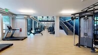 This two-story fitness center in Salt Lake City, Utah, offers a well-equipped and thoughtfully designed exercise environment spread across a generously sized main floor and a smaller upper-level studio. The main floor is the heart of the facility, featuring soaring floor-to-ceiling windows along one wall that flood the space with natural light and provide outdoor views of surrounding trees and the cityscape. The open layout accommodates a wide range of equipment, including treadmills, rowing machines, ellipticals, cable machines, and a multi-functional Life Fitness functional trainer. Battle ropes and weight plates are also available for functional training. Exposed industrial-style ductwork runs along the upper sections of the walls, adding a contemporary aesthetic that complements the clean white columns and light hardwood-style flooring throughout. A bold navy blue accent wall anchors the staircase area, where a wall-mounted display screen provides an engaging focal point. A glass-paneled room offers a semi-private training area, while open built-in shelving provides organized storage. The striking dark metal staircase leads to the upper level, which features a mirrored barre wall, stationary cycling bikes, and additional workout space—ideal for focused, small-group sessions or individual training.