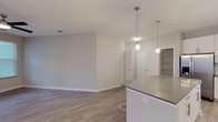 This single-floor residence at 2599 Evans Rd in Morrisville, NC offers a clean, modern aesthetic with thoughtfully designed living spaces and quality finishes throughout. The open-concept layout seamlessly connects the kitchen and living areas, creating a bright and functional flow. The kitchen is a standout feature, equipped with stainless steel appliances including a refrigerator with ice maker, electric range, microwave, and dishwasher. White shaker-style cabinetry extends to the ceiling for ample storage, complemented by a subway tile backsplash, dark quartz countertops, and a large center island with pendant lighting above. Light wood-look flooring runs throughout the main living areas, adding warmth and continuity. The living room is generously sized, featuring a ceiling fan, access to an outdoor area, and windows with blinds that invite natural light. Bedrooms are carpeted for comfort, with one featuring a walk-in closet outfitted with wire shelving. The bathrooms are well-finished — one includes a soaking tub with tile surround, while another offers a glass-door walk-in shower, both with wood-look flooring, gray quartz vanity tops, and white cabinetry. Additional built-in shelving provides convenient storage throughout the unit.