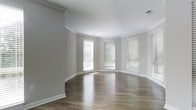 This single-floor space showcases a thoughtfully designed interior that blends classic architectural details with a clean, modern aesthetic. The standout feature of the main living area is its gracefully curved wall, which lends a distinctive and sophisticated character to the room. Warm hardwood flooring runs throughout, reflecting light and adding a sense of warmth and continuity to the space.

Crown molding lines the ceilings, adding a refined finishing touch that enhances the overall elegance of the interior. The walls are finished in a soft, neutral gray tone that creates a calm and versatile backdrop, complemented by crisp white baseboards and trim throughout.

Natural light fills the space generously through multiple windows fitted with horizontal blinds, offering both brightness and privacy control. The windows are positioned along the curved wall, amplifying the architectural interest of the room while framing views of the surrounding greenery outside.

The overall ambiance is open, airy, and inviting — a well-lit, move-in-ready space that offers a neutral canvas ideal for a variety of furnishing styles. The combination of classic detailing, quality flooring, and abundant natural light makes this a comfortable and visually appealing environment.