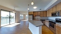 This single-floor residence offers a thoughtfully designed open-concept layout that seamlessly connects the kitchen and living areas into one cohesive, flowing space. The kitchen is well-appointed with warm honey-toned wood cabinetry, gray countertops, and stainless steel appliances, including a refrigerator and dishwasher. A central kitchen island with a gray countertop provides additional prep space and serves as a natural divider between the kitchen and the rest of the living area. Pendant lighting fixtures hang above the island, adding both function and style to the space. Durable hardwood flooring runs throughout, tying the entire space together with a warm, consistent aesthetic. The living area is generously sized, bathed in natural light streaming through multiple windows, and features a ceiling fan for added comfort. A sliding glass door leads to an outdoor area, further enhancing the sense of openness and bringing in additional light. The overall ambiance is clean, modern, and inviting, with a neutral color palette that creates a versatile backdrop for a variety of interior styles.