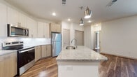 This single-floor residence offers a thoughtfully designed layout with a contemporary aesthetic carried consistently throughout its spaces. The kitchen serves as the centerpiece, showcasing a large granite-topped island with an undermount sink and stylish pendant lighting overhead. Cabinetry combines crisp white upper cabinets with warm natural wood lower cabinets, complemented by a white subway tile backsplash and a full suite of stainless steel appliances, including a refrigerator, range, and microwave. Wood-look plank flooring runs through the kitchen and main living areas, adding warmth and durability.

The living area is bright and open, featuring a ceiling fan, vertical blinds, and direct access to adjoining rooms. Bedrooms are finished with plush carpet, ceiling fans, and window blinds that allow natural light while maintaining privacy.

The bathroom features large-format beige tile surrounding a soaking tub and shower combination, with brushed bronze fixtures adding a refined touch. A separate vanity area includes granite countertops and wood-toned cabinetry. An in-unit laundry area with washer and dryer hookups provides added convenience. Overall, the space presents a clean, modern aesthetic with practical, well-appointed finishes.