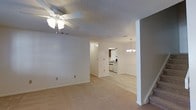 Located at 10875 Abercorn St in Savannah, Georgia, this two-floor space offers a practical and well-organized layout designed for comfortable living. The main level features an open living area with neutral-toned carpeting and light-colored walls that create a bright, airy atmosphere throughout. A ceiling fan with a light fixture is centrally positioned overhead, promoting air circulation and adding a functional accent to the room. A structural column subtly defines the transition between the living and dining areas, while a chandelier pendant light illuminates the dining space with a classic touch. The kitchen is visible from the main living area and is equipped with white cabinetry, standard appliances, and tile flooring, offering an efficient workspace. A textured popcorn ceiling is consistent throughout the main level, and white baseboards frame each room cleanly. Stairs lead to the second floor, which provides additional living space above. The overall aesthetic is clean and understated, with a neutral color palette that lends versatility to the interior. The layout facilitates easy movement between connected areas, making the space functional and adaptable for a variety of uses.